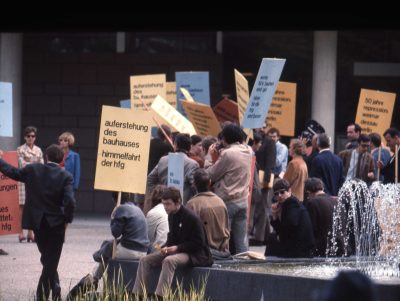Personengruppe in Rückansicht vor einem Brunnen stehend und mit Demonstrationsschildern in den Händen.